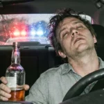 Drunk driver holding alcohol bottle with police lights flashing behind car.