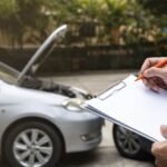 A close-up of a person's hands writing on a clipboard with an orange pen, with a blurred silver car with its hood open in the background.