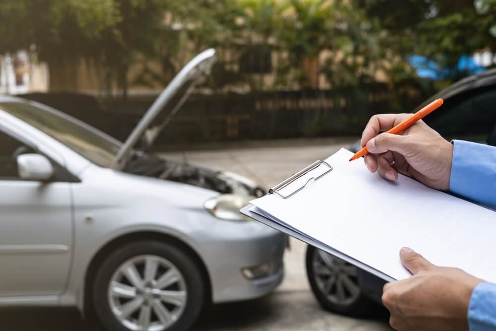 A close-up of a person's hands writing on a clipboard with an orange pen, with a blurred silver car with its hood open in the background.