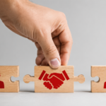 Hand placing a puzzle piece with red partnership icons between two wooden person-icon pieces.
