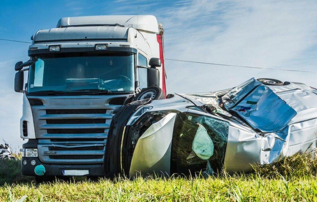 A semi-truck parked next to a severely damaged, overturned car on the side of a grassy road.