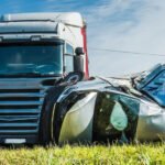 A semi-truck parked next to a severely damaged, overturned car on the side of a grassy road.