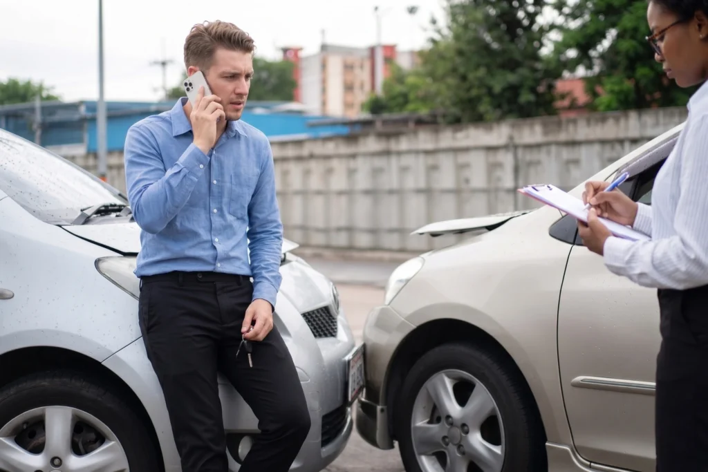 A man talking on his phone next to a minor car accident while a woman takes notes on a clipboard.