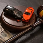 Gavel beside crashed toy cars and dollar bills on a dark table.