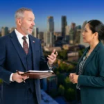 Two professionals in business suits are having a conversation with a blurred city skyline background.
