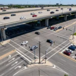 High-angle view of a multi-lane city intersection with traffic, situated directly beneath a large, elevated concrete highway overpass carrying several cars.