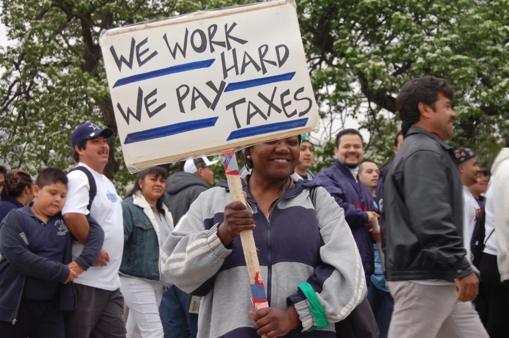 A smiling woman holding a handwritten sign that reads "WE WORK HARD, WE PAY TAXES" while marching in a crowd of people outdoors.