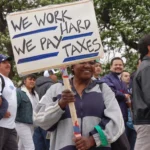 A smiling woman holding a handwritten sign that reads "WE WORK HARD, WE PAY TAXES" while marching in a crowd of people outdoors.