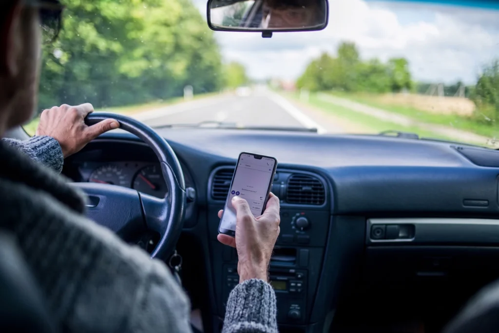 View from inside a moving car showing a driver holding a smartphone in one hand while steering with the other along a two-lane road.