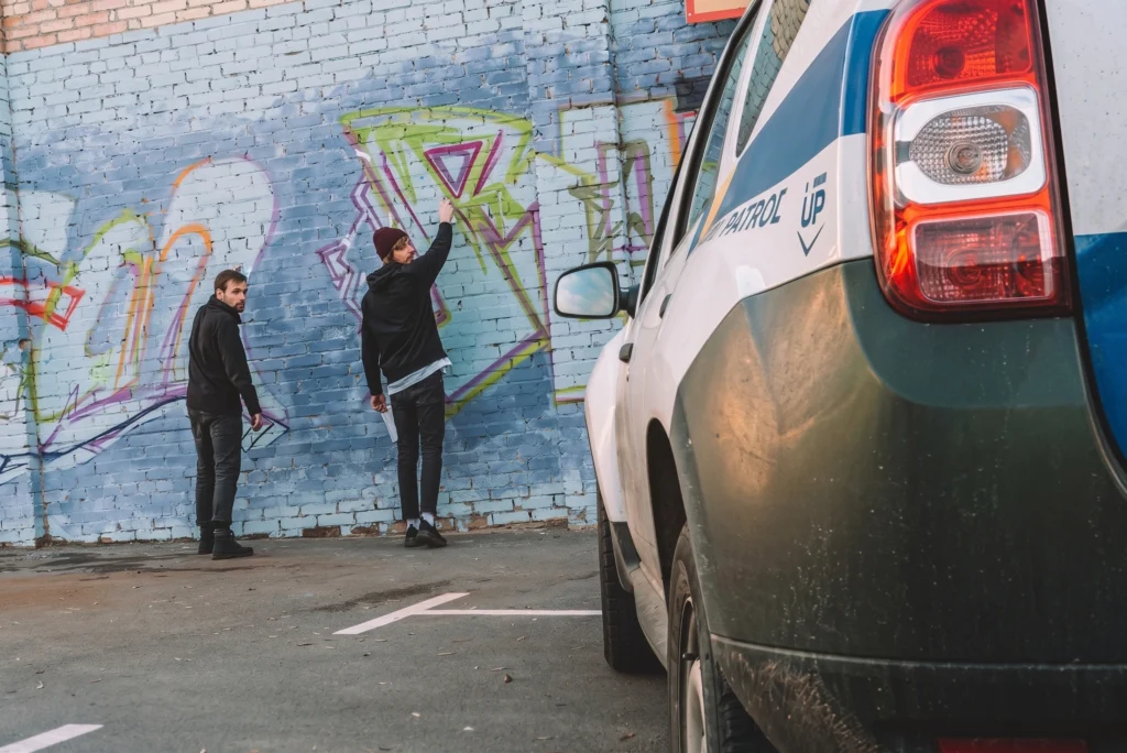 Two young men spray-painting graffiti on a brick wall as a police vehicle approaches in the foreground.