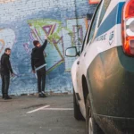 Two young men spray-painting graffiti on a brick wall as a police vehicle approaches in the foreground.