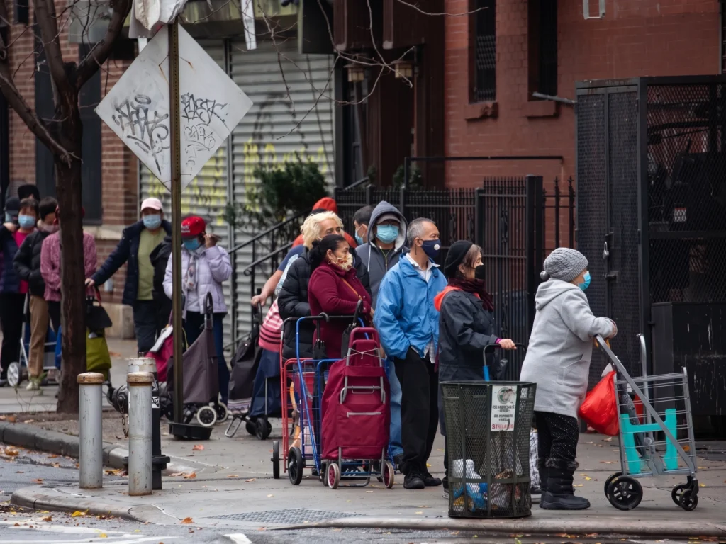 A line of people wearing face masks and holding shopping carts queue along a city sidewalk.