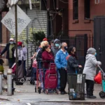 A line of people wearing face masks and holding shopping carts queue along a city sidewalk.