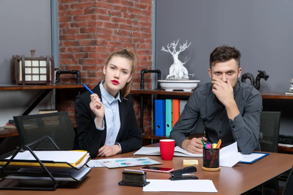 Two office professionals seated at a desk looking serious and concerned, with documents and office supplies in front of them