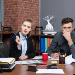 Two office professionals seated at a desk looking serious and concerned, with documents and office supplies in front of them