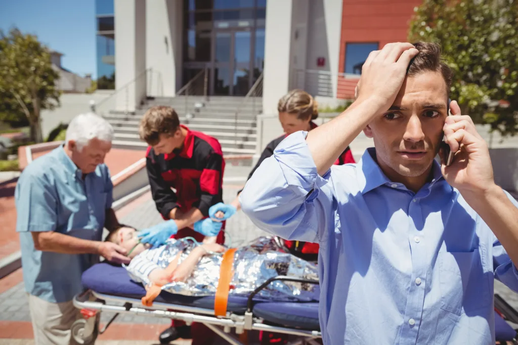 A distressed man in a blue shirt talking on a phone call while holding his head, with paramedics treating an injured person on a stretcher in the background.
